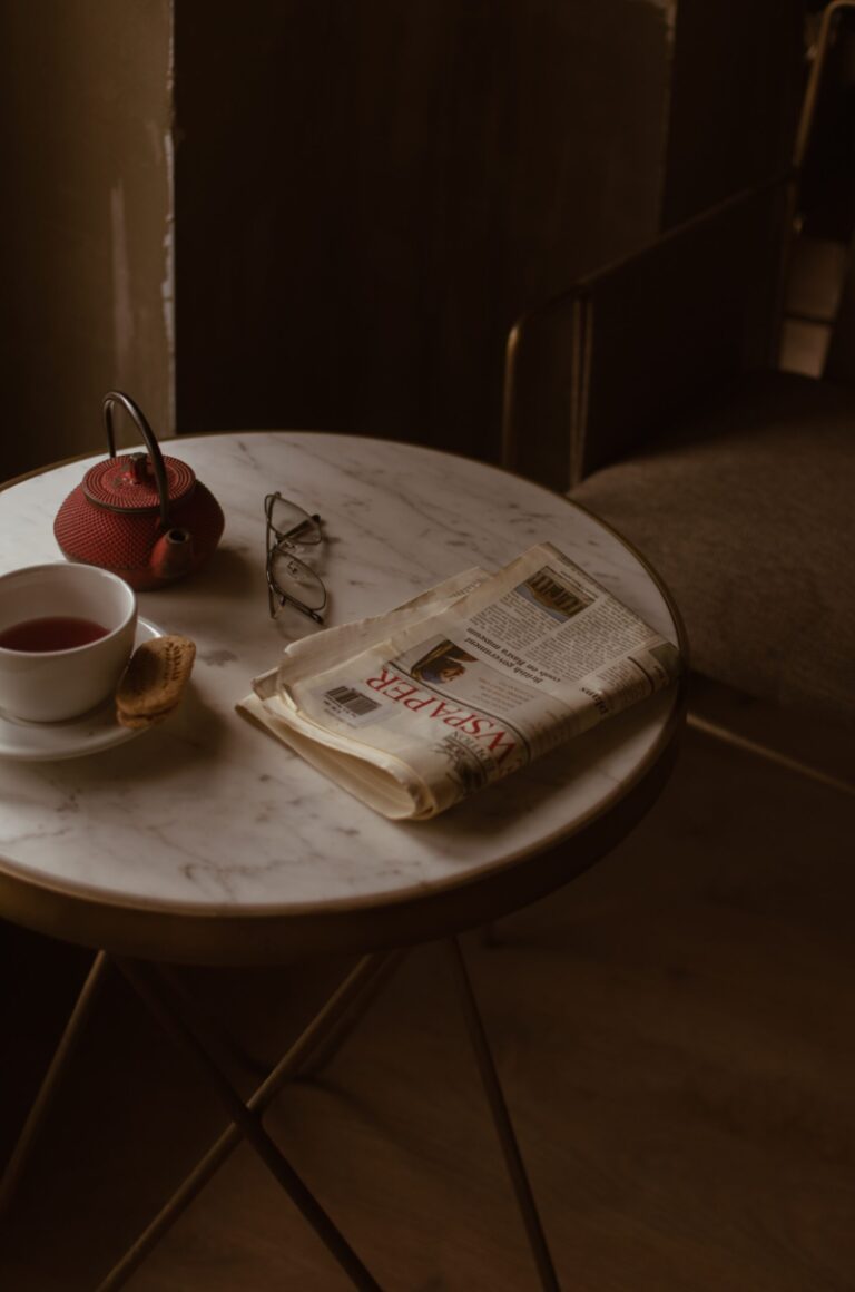 newspaper on a marble table with reading glasses, a tea cup, and a teapot